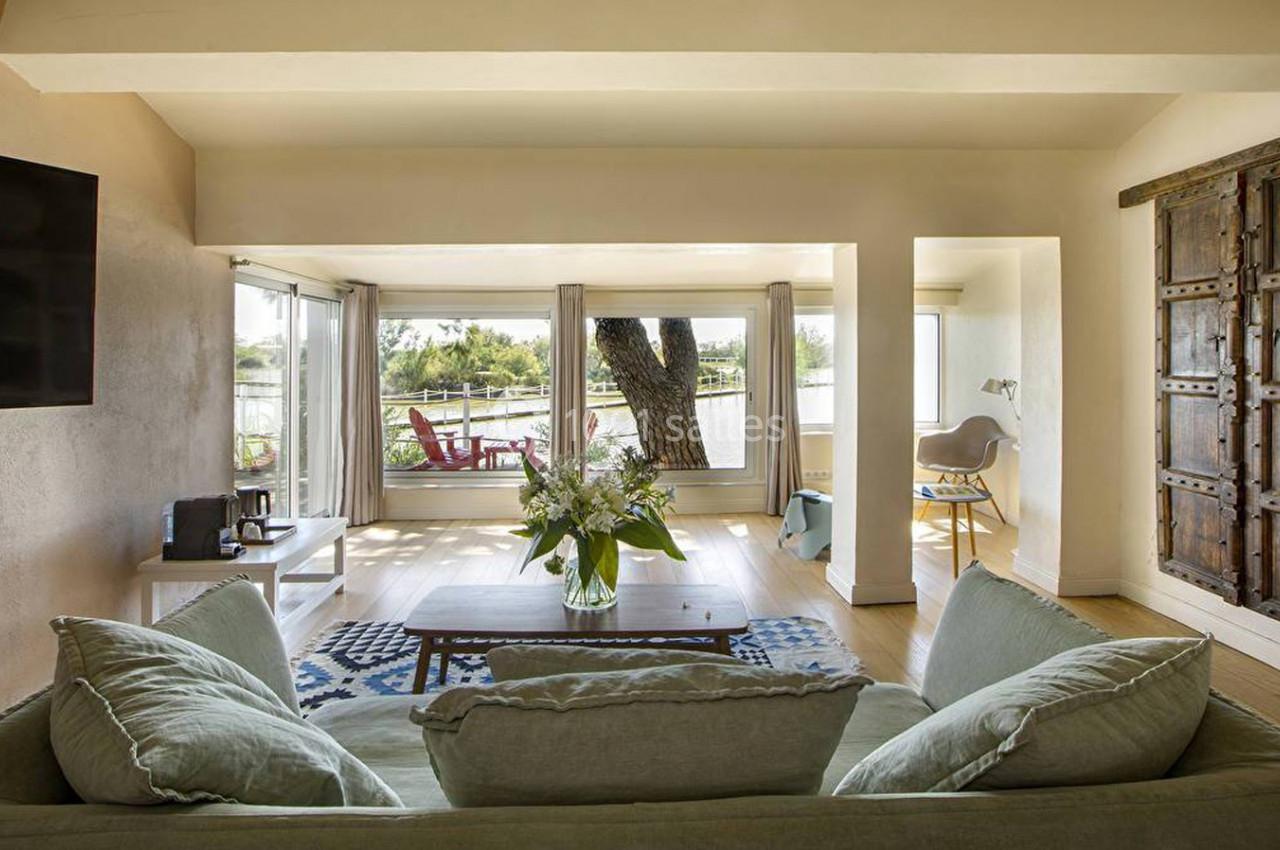 Salon lumineux avec canapé vert, table basse, grandes fenêtres donnant sur une terrasse avec vue sur un paysage naturel.