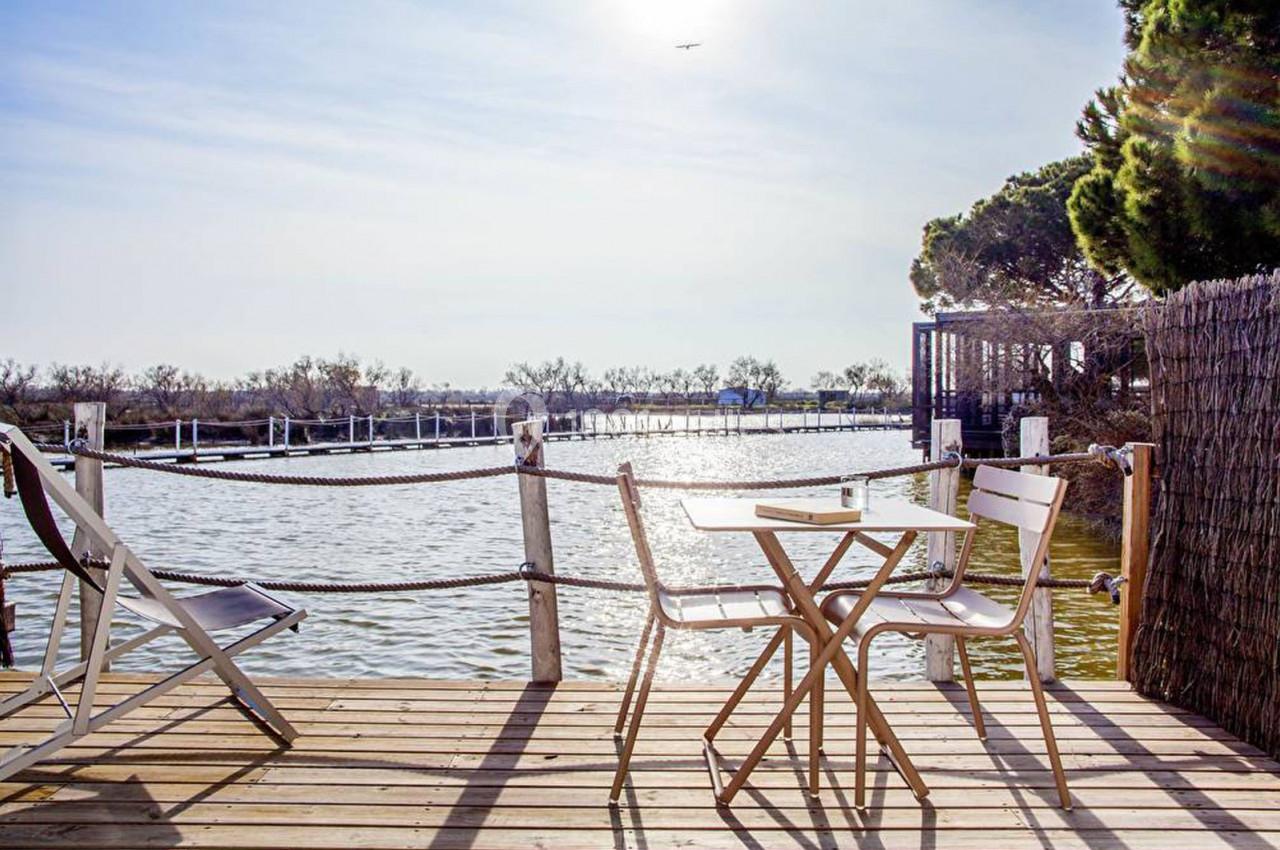 Terrasse en bois avec table et chaises blanches donnant sur un plan d'eau entouré de végétation et de clôtures en bois.