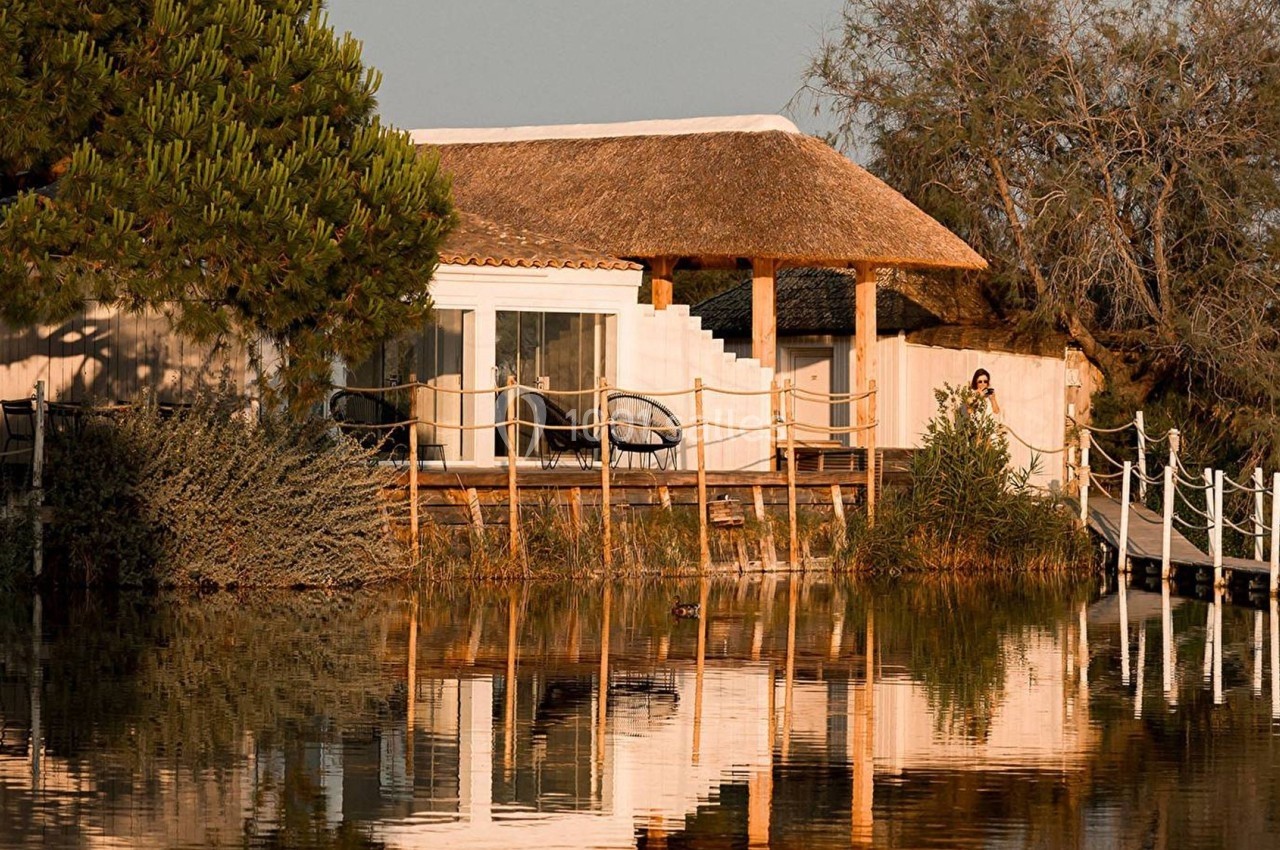 Maison au toit de chaume bordant un étang, entourée de végétation et accessible par une passerelle en bois.