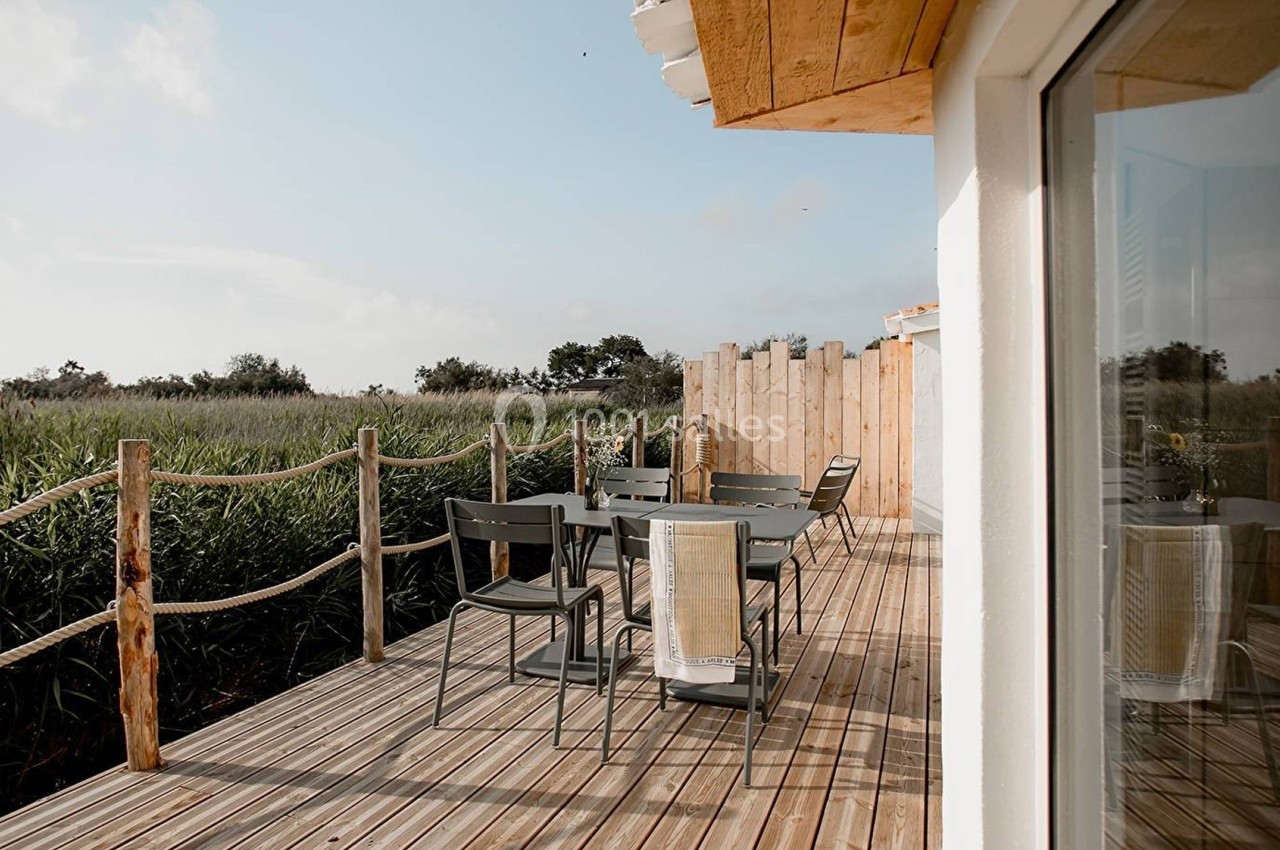 Terrasse en bois avec table et chaises, entourée de végétation et clôturée par des cordes, sous un ciel dégagé.
