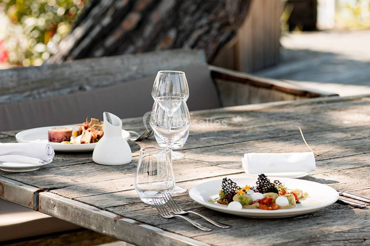 Table en bois avec deux assiettes de plats gastronomiques, verres et serviettes, en extérieur sous un arbre.