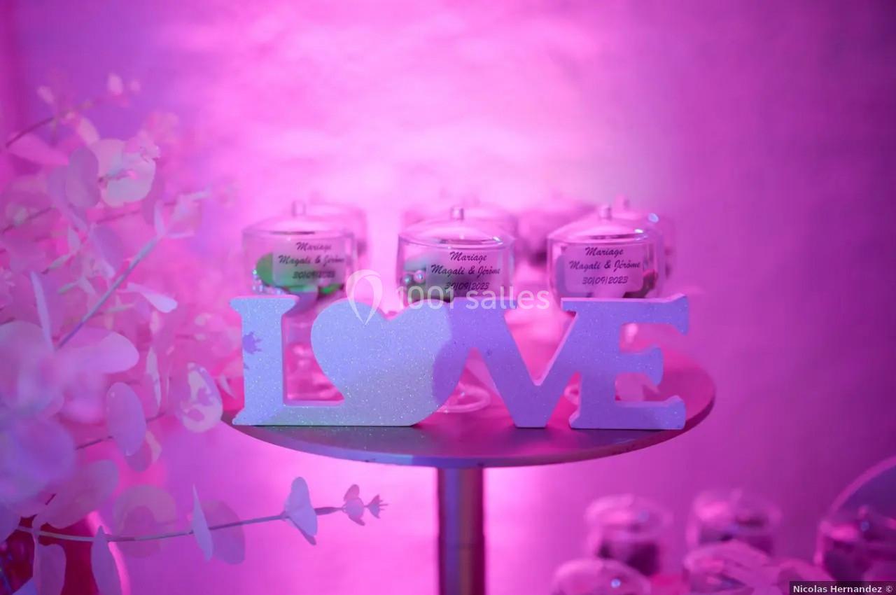 Décoration rose avec le mot ’LOVE’ en lettres découpées, posée sur une table devant des bougies en arrière-plan.