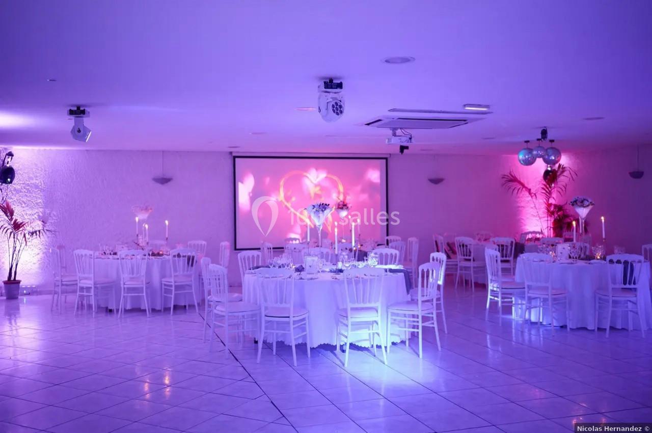 Salle de réception décorée avec des tables rondes, des chaises blanches et un éclairage rose-violet tamisé.