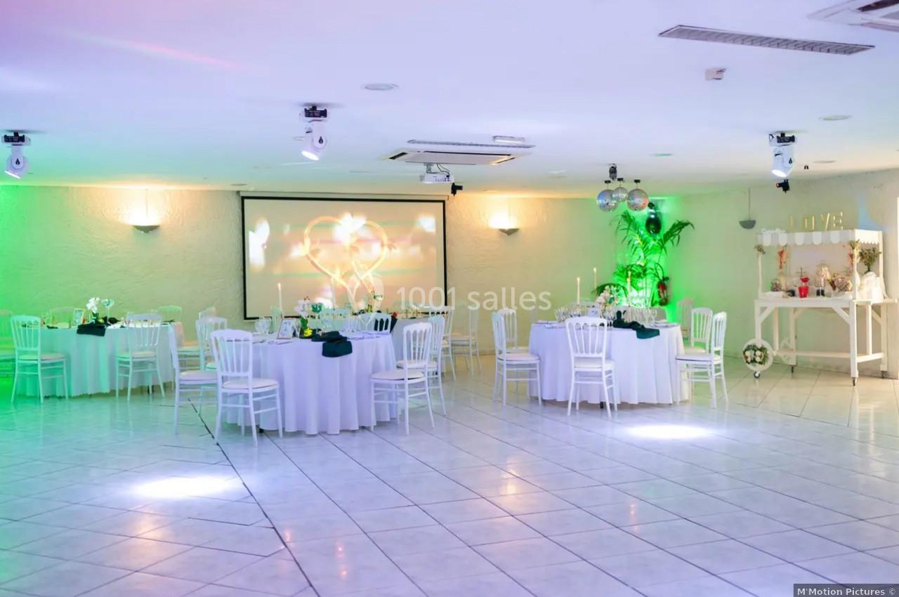 Salle de réception lumineuse avec tables rondes décorées, chaises blanches et éclairage vert.