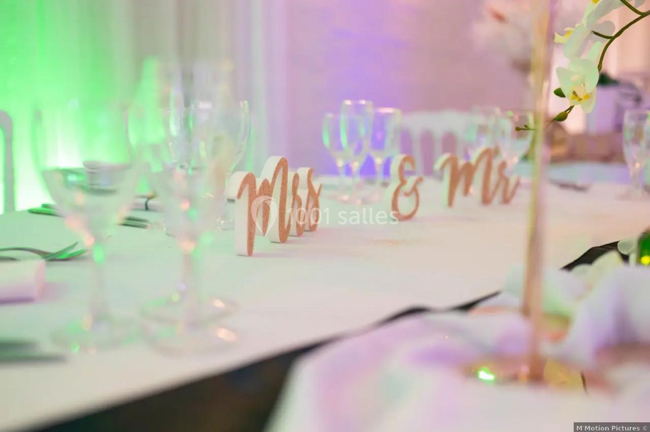 Décoration de table de mariage avec des marqueurs ’Mrs & Mrs’, verres à vin et éclairage coloré en arrière-plan.