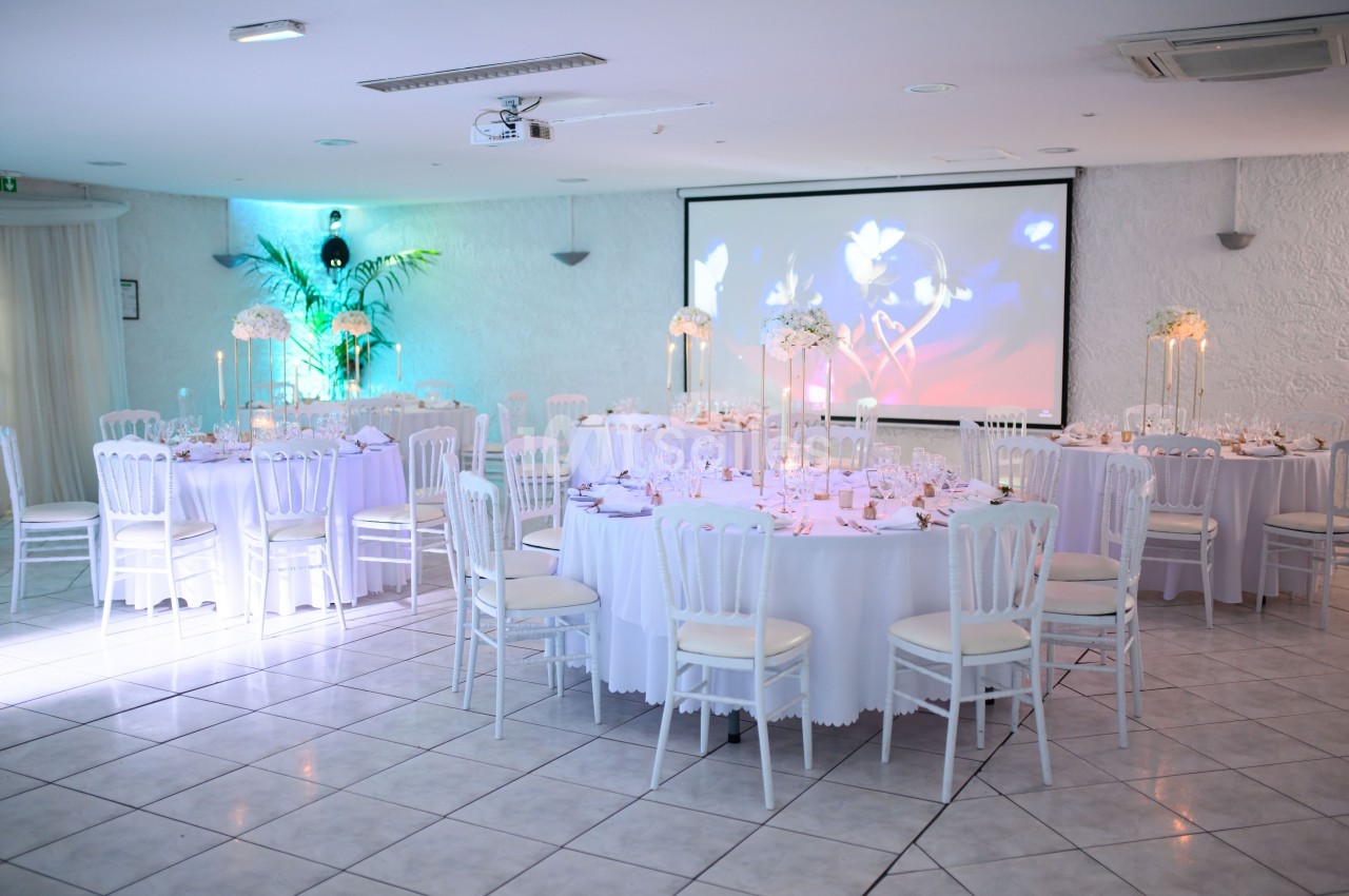 Salle de réception décorée avec des tables rondes, nappes blanches, chandeliers et fleurs, éclairage tamisé.