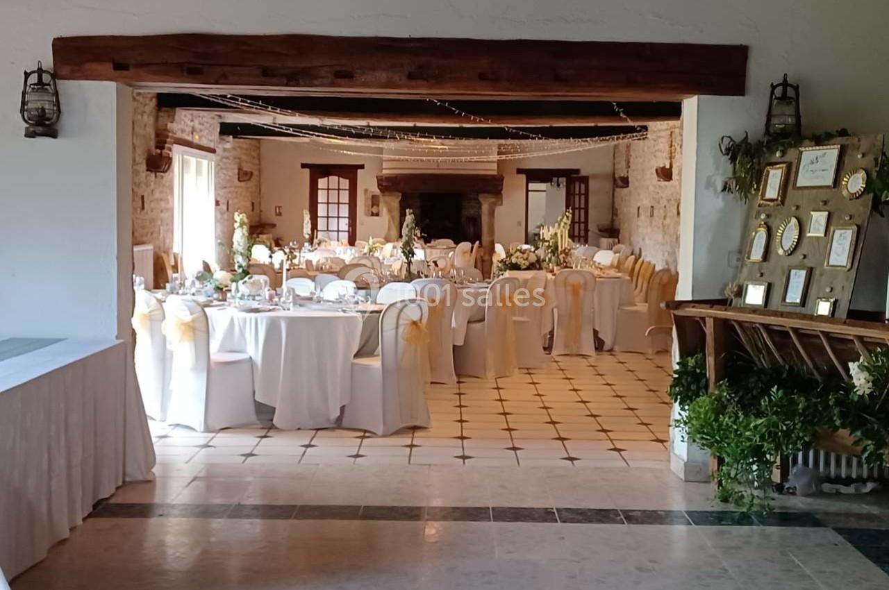 Salle de réception décorée avec des tables rondes, nappes blanches, chaises ornées de rubans dorés et cadres exposés.