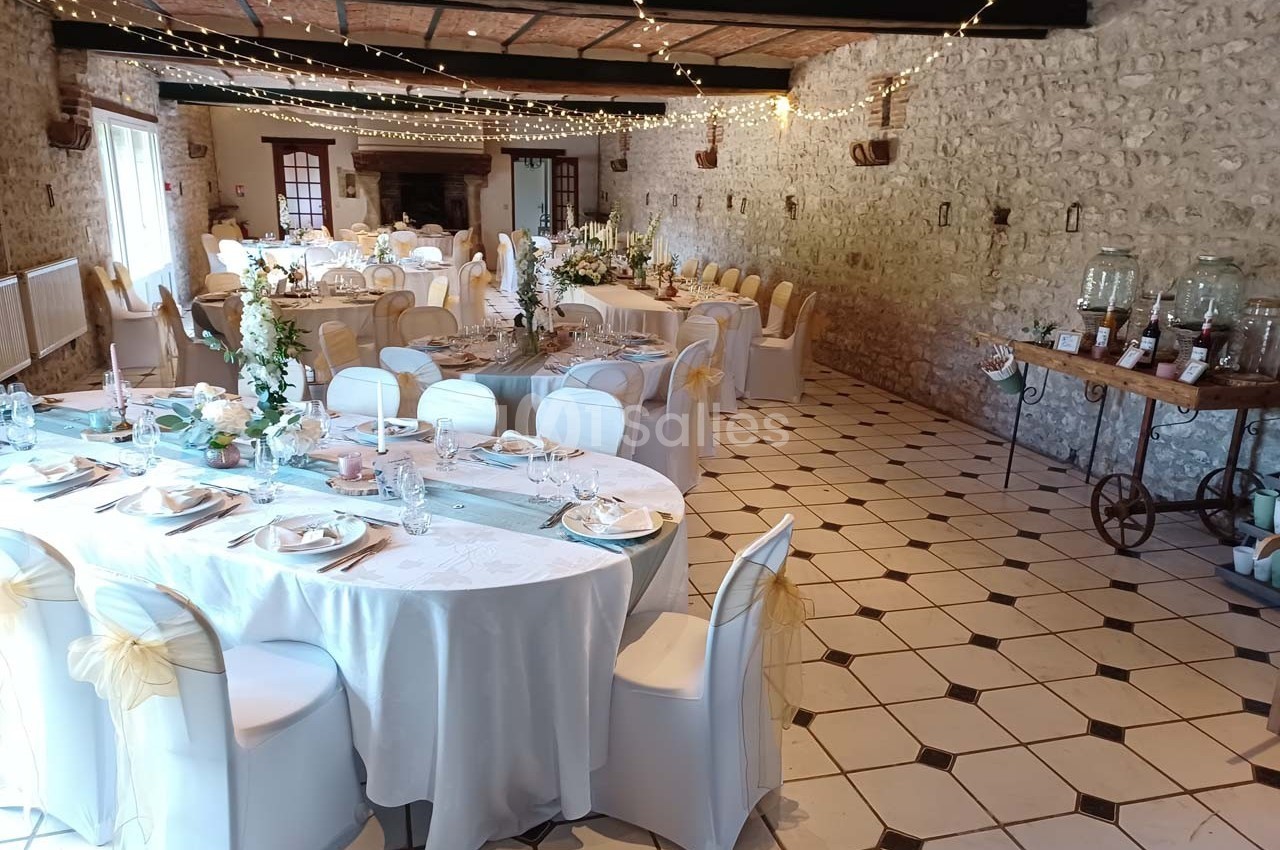 Salle de réception décorée pour un mariage, avec tables dressées, chaises ornées de nœuds et guirlandes lumineuses…