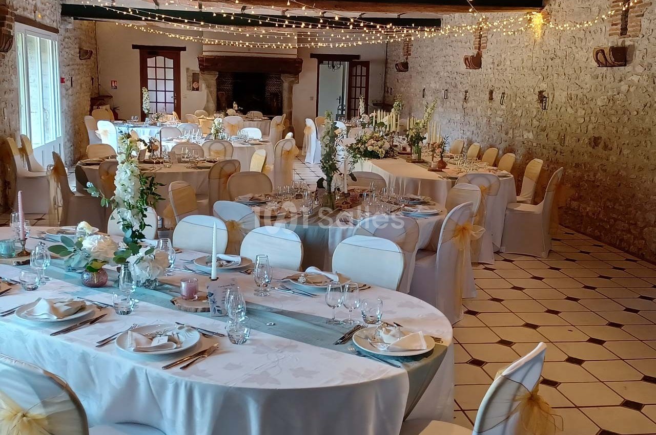 Salle de réception décorée pour un mariage, avec tables dressées, chaises ornées de rubans et guirlandes lumineuses…