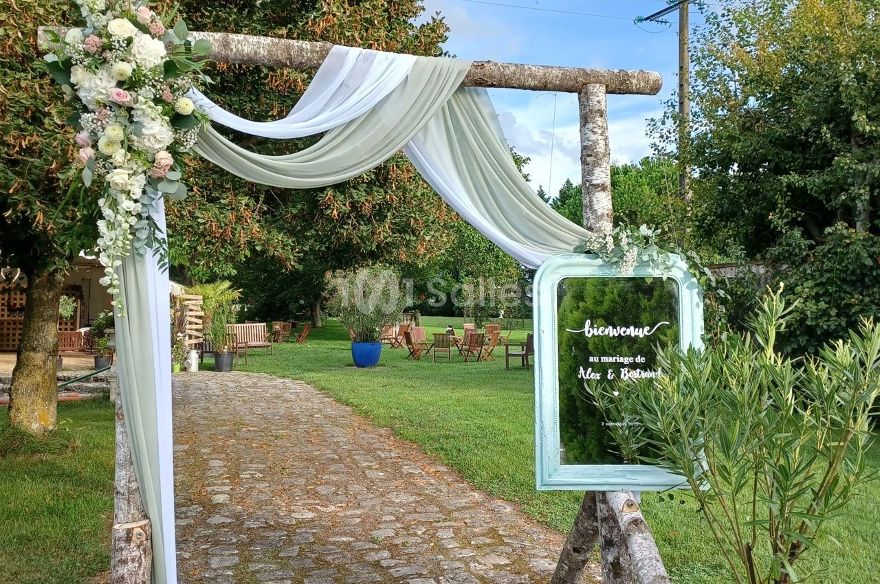 Arche en bois décorée de fleurs et de voilages, avec un miroir indiquant un mariage, dans un jardin verdoyant.