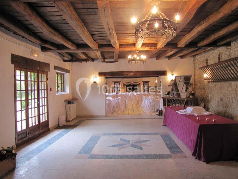 Salle de réception avec poutres apparentes, tables dressées au fond et buffet couvert d'une nappe rouge à droite.