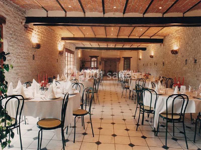 Salle de réception avec murs en pierre, tables rondes dressées et chaises noires, éclairée par des appliques murales.