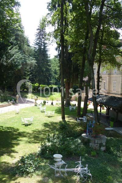 Jardin verdoyant avec tables blanches dispersées, entouré d'arbres et jouxtant un bâtiment ancien en pierre.