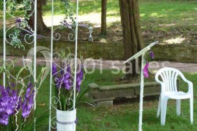 Deux fauteuils en rotin décorés, placés sous une arche ornée de fleurs et de tissus, dans un jardin verdoyant.