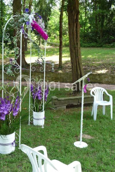 Arche décorée de fleurs violettes et blanches avec chaises en plastique blanc dans un jardin verdoyant.