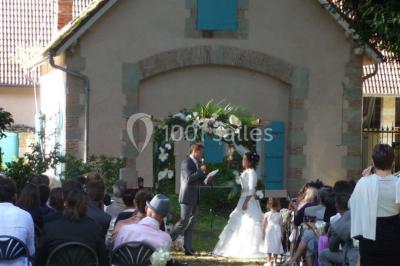 Deux fauteuils en rotin décorés, placés sous une arche ornée de fleurs et de tissus, dans un jardin verdoyant.