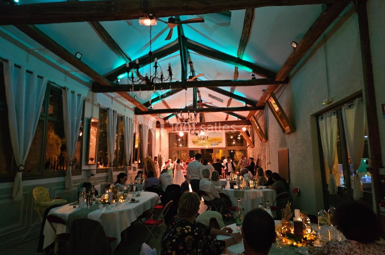Salle de réception éclairée en soirée, avec des invités assis à des tables décorées et une piste de danse au fond.