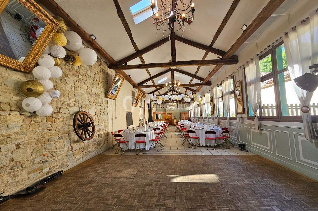 Salle de réception lumineuse avec poutres apparentes, tables dressées, décorations suspendues et murs en pierre.