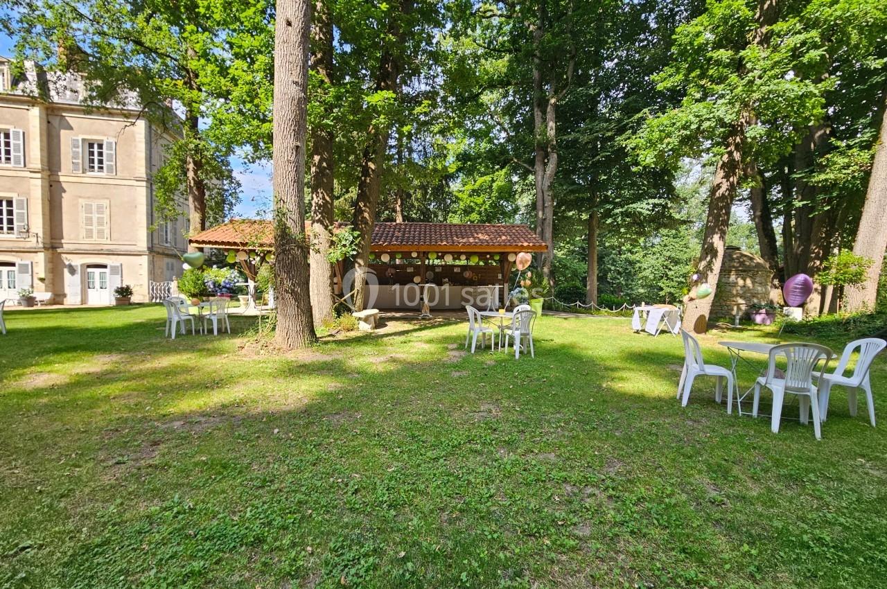 Espace extérieur avec pelouse, tables et chaises blanches, cabanon décoré et arbres entourant un bâtiment ancien.