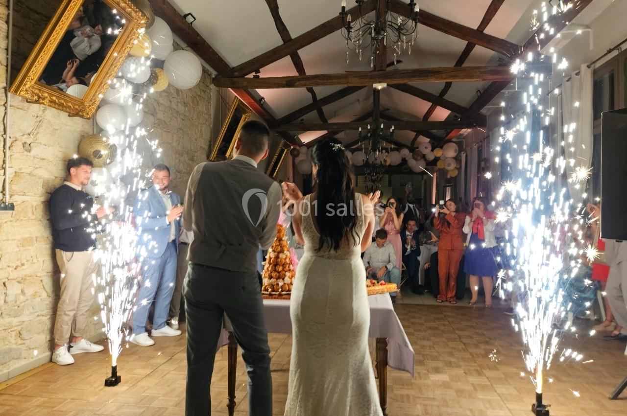 Un couple de mariés coupe une pièce montée dans une salle décorée, entouré d'invités et de fontaines lumineuses.