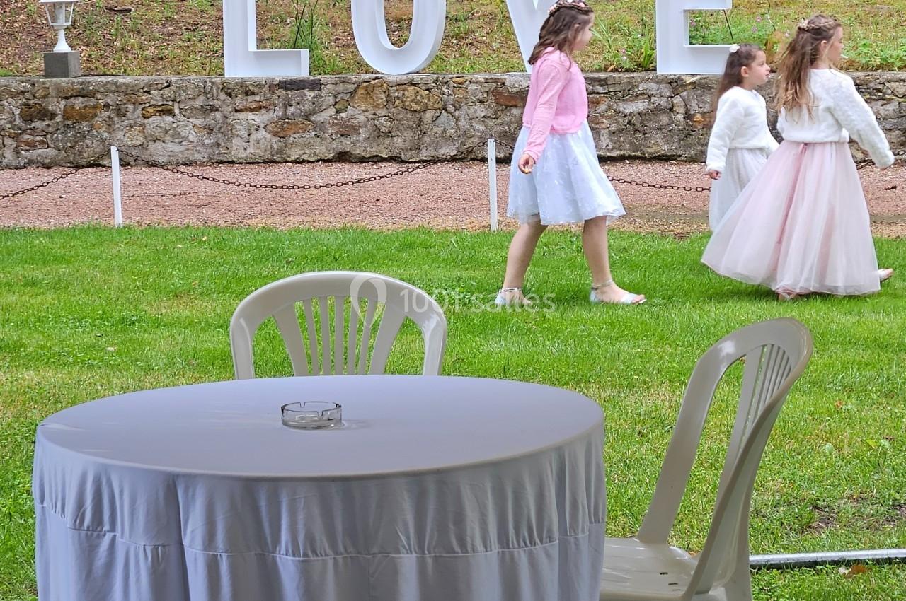 Table ronde avec nappe blanche et chaises en plastique sur pelouse, enfants en tenue élégante marchant près d'un panneau…