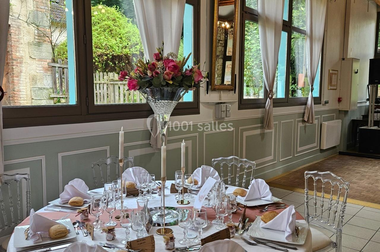 Table élégamment dressée avec nappes blanches, vaisselle raffinée, bougies et bouquet de fleurs, dans une salle lumineuse.