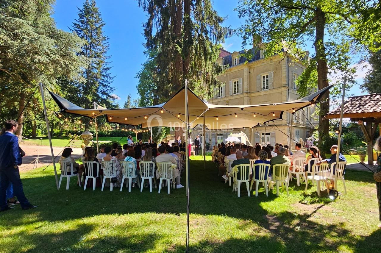 Cérémonie en plein air sous une tente, avec des invités assis sur des chaises blanches, devant un bâtiment ancien.
