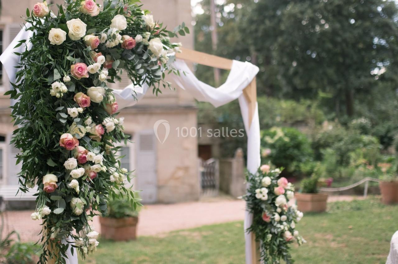 Arche de mariage en bois décorée de fleurs blanches et roses, entourée d'un jardin verdoyant.