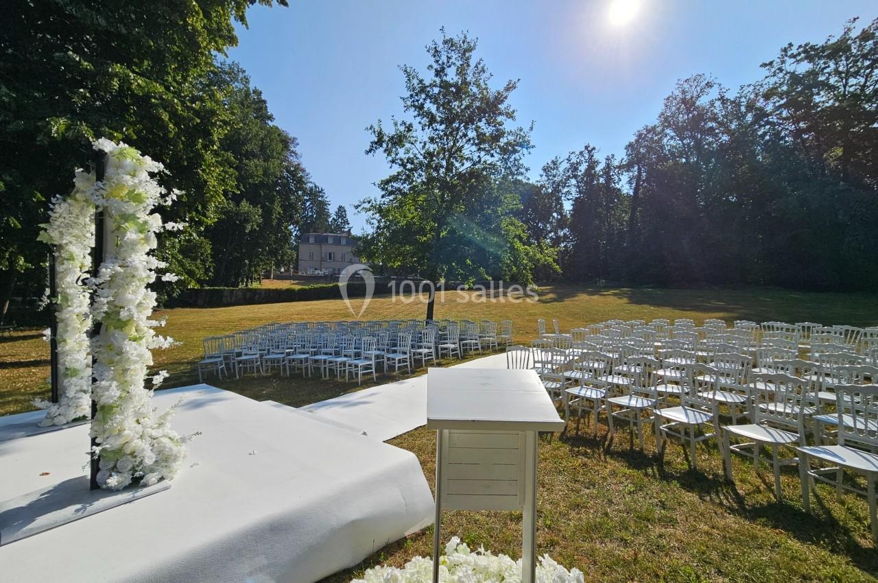 Cérémonie en plein air avec chaises blanches alignées, arche fleurie et pupitre, sous un ciel ensoleillé.