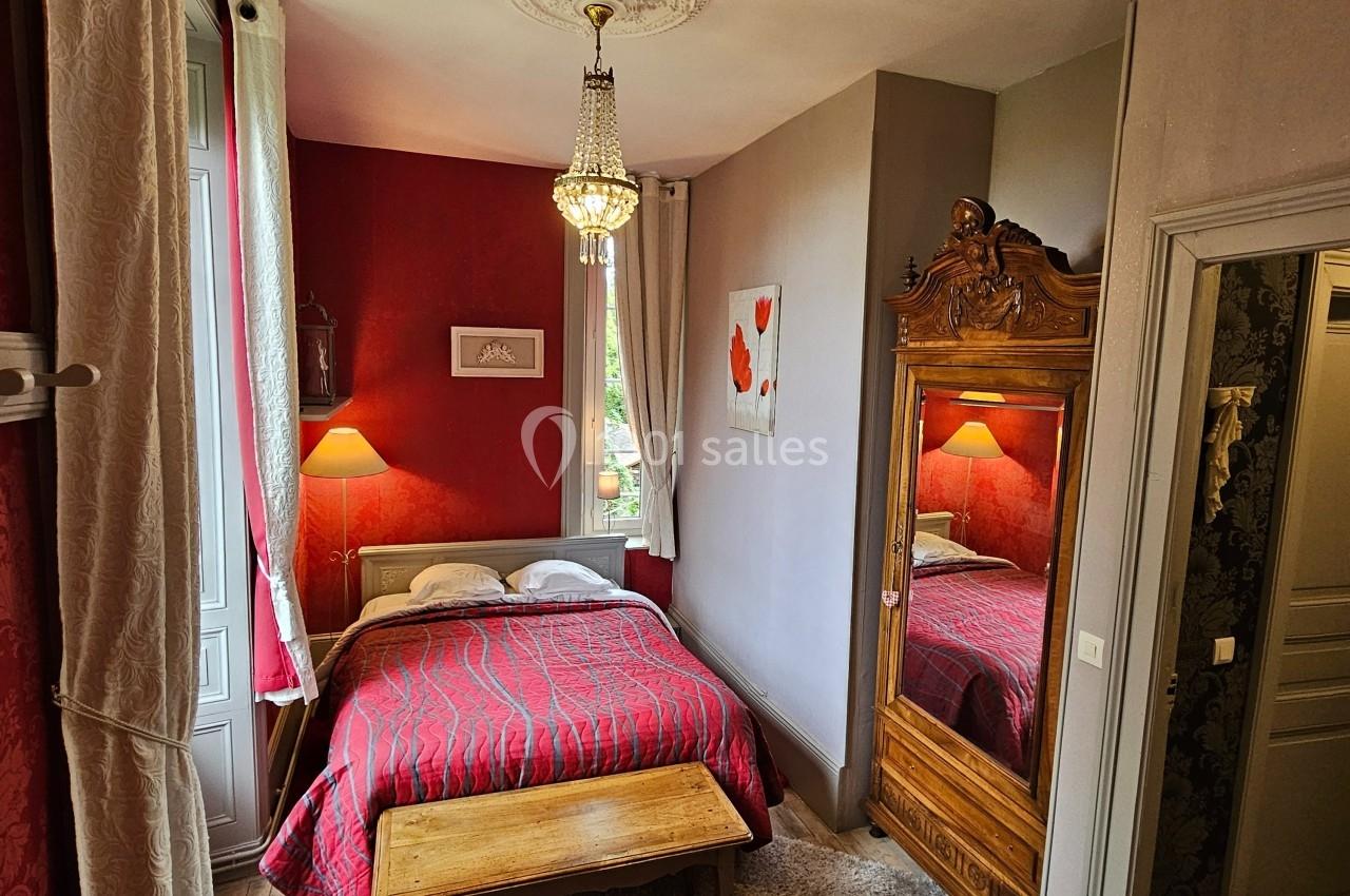 Chambre avec lit double, armoire en bois sculpté, lustre, murs rouges et gris, et tapis sur parquet clair.