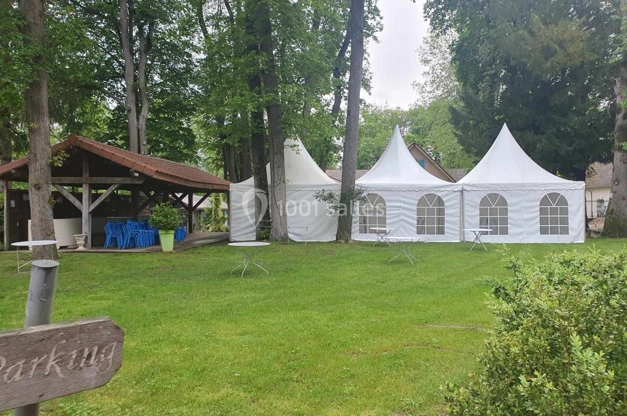 Tentes blanches et abri en bois installés dans un espace vert entouré d'arbres, avec des tables disposées sur la pelouse.