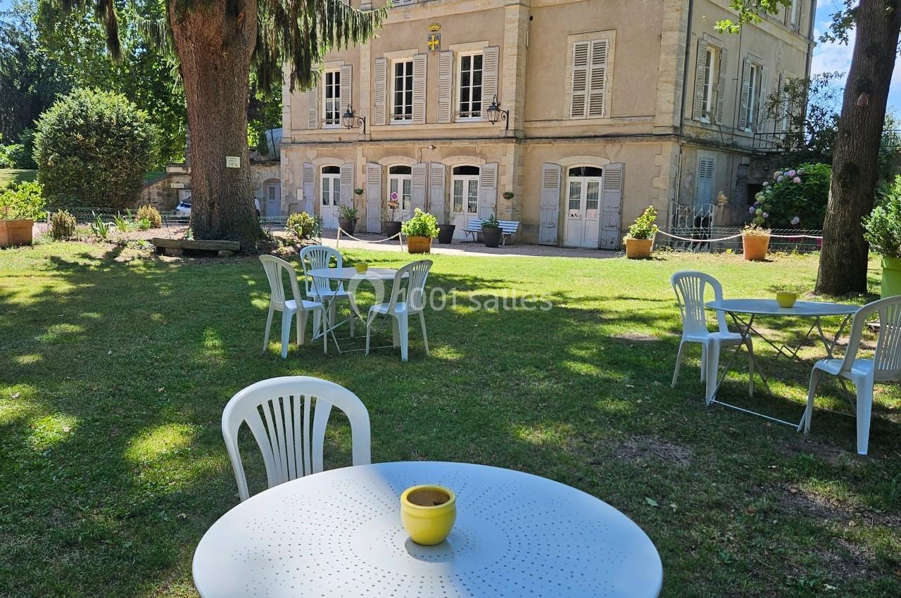 Tables et chaises blanches disposées dans un jardin ombragé devant un grand bâtiment ancien en pierre.
