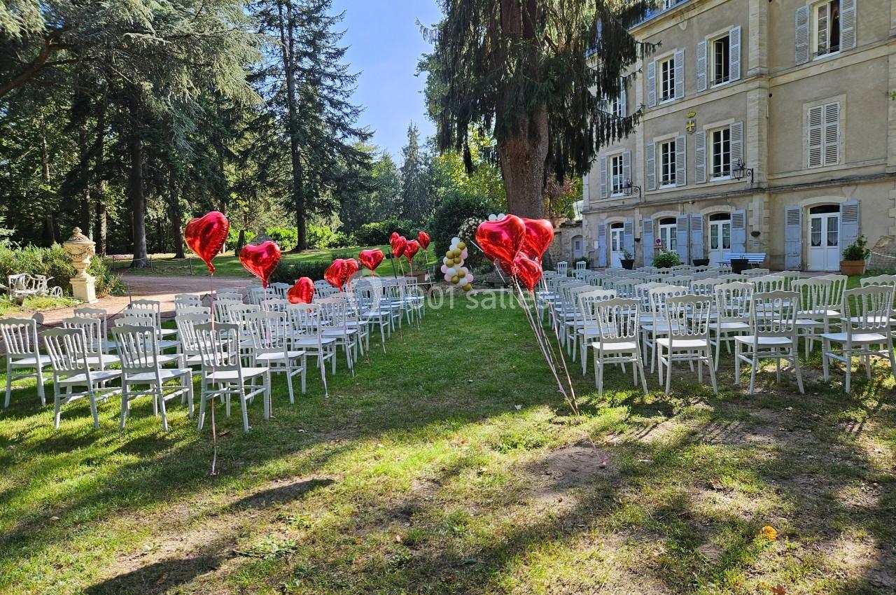 Chaises blanches alignées en extérieur pour une cérémonie, avec des ballons rouges en forme de cœur et un bâtiment en…