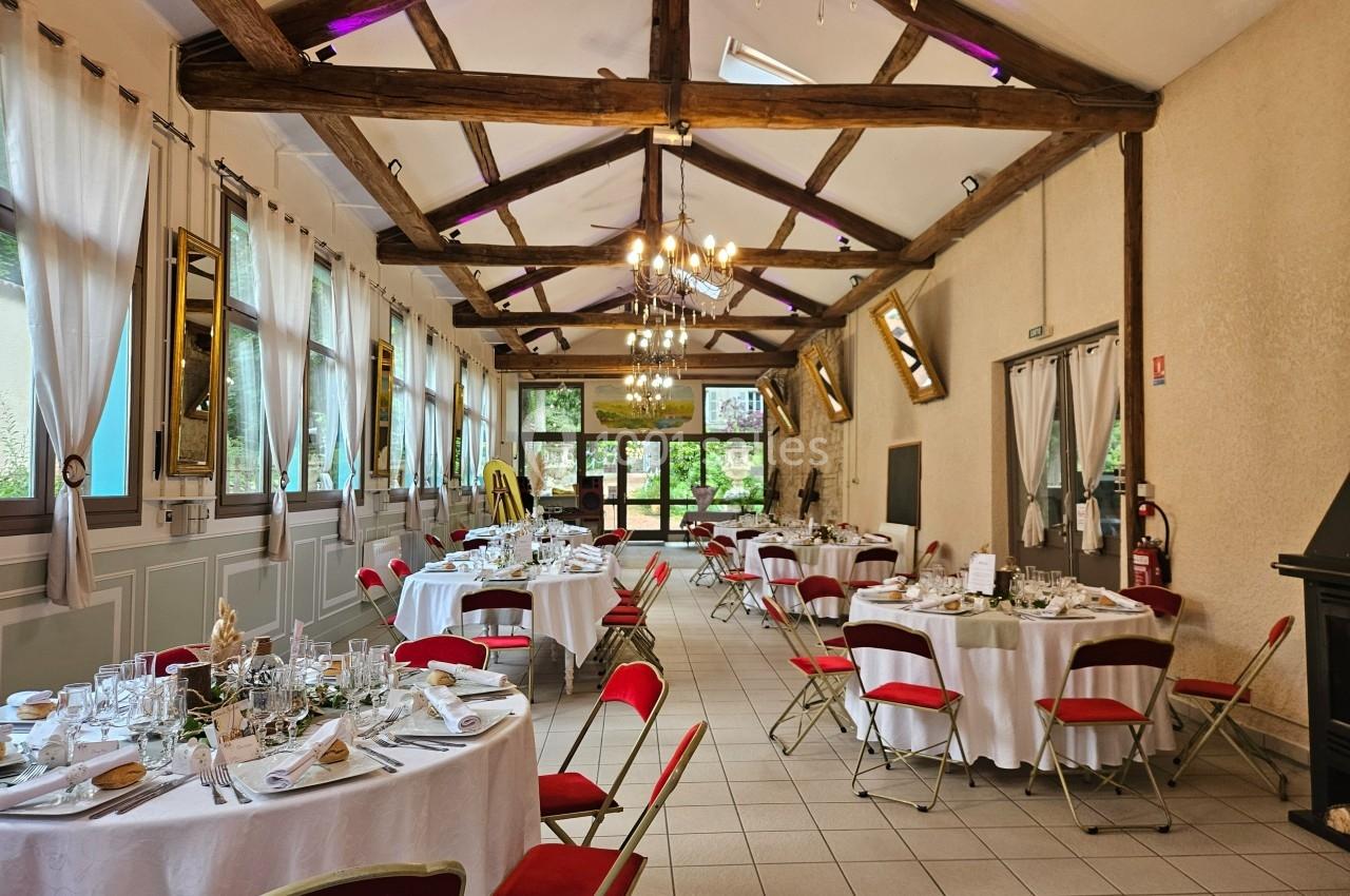 Salle de réception lumineuse avec poutres apparentes, tables rondes dressées et chaises rouges.