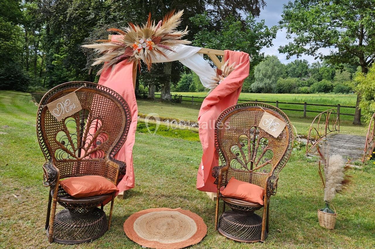 Deux fauteuils en rotin décorés, placés sous une arche ornée de fleurs et de tissus, dans un jardin verdoyant.