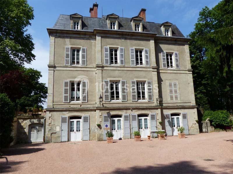 Façade d'un bâtiment ancien à trois étages avec volets blancs, entouré de végétation et d'une cour en gravier.