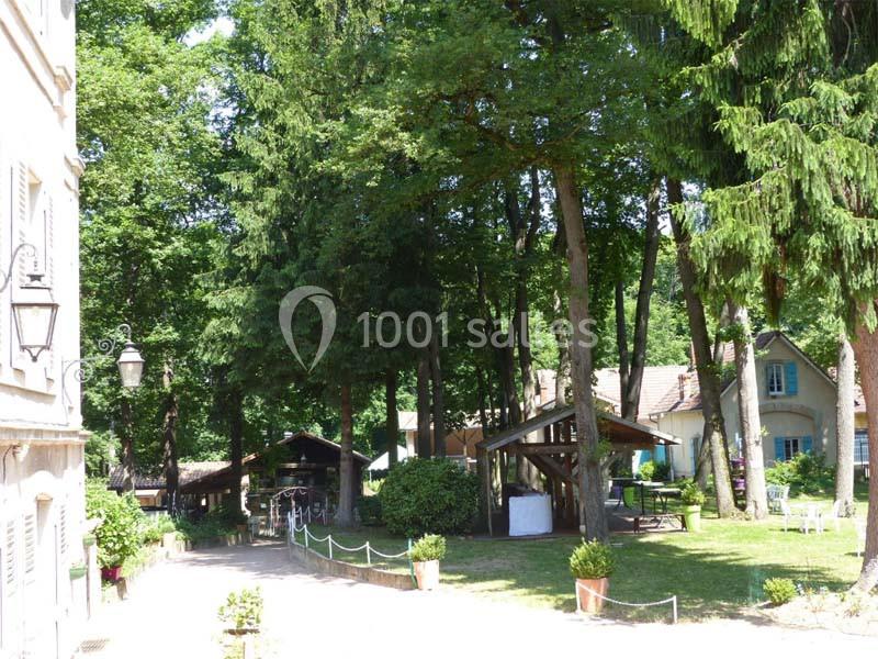 Cour ombragée avec des arbres, des bâtiments en arrière-plan et des tables disposées sous des abris en bois.