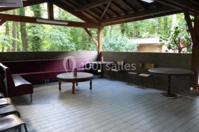 Espace couvert avec canapé, tables et chaises en bois, entouré de verdure visible à travers les ouvertures.