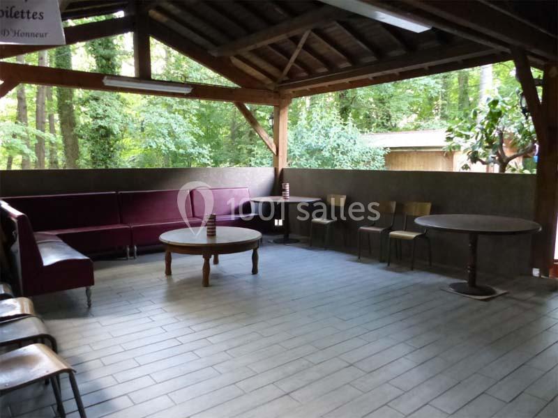 Espace couvert avec canapé, tables et chaises en bois, entouré de verdure visible à travers les ouvertures.