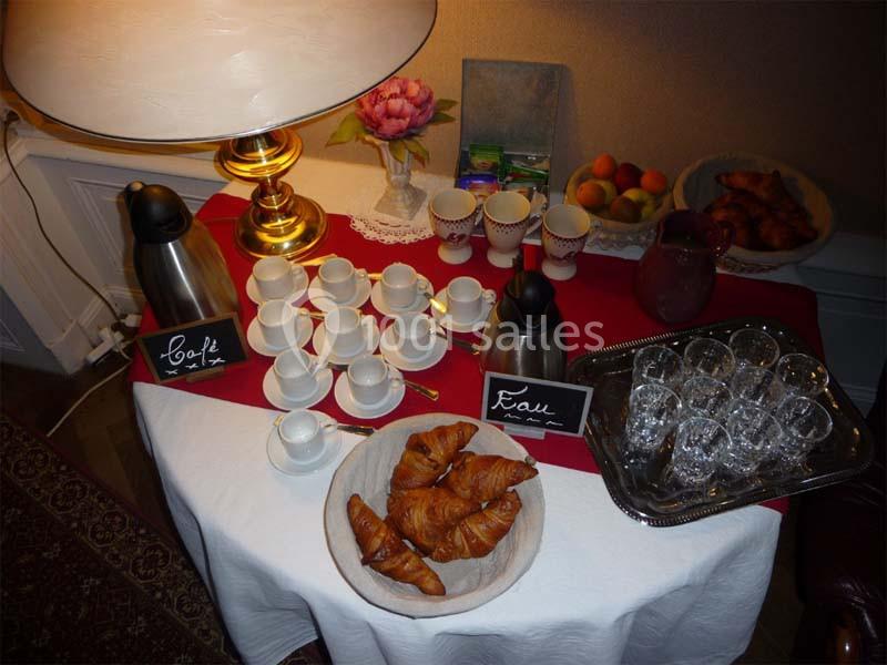 Table avec cafetière, carafe d'eau, tasses, croissants, fruits et verres, éclairée par une lampe de table.