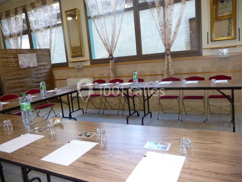Salle de réunion avec tables disposées en U, chaises rouges, documents, bouteilles d'eau et fenêtres avec rideaux.