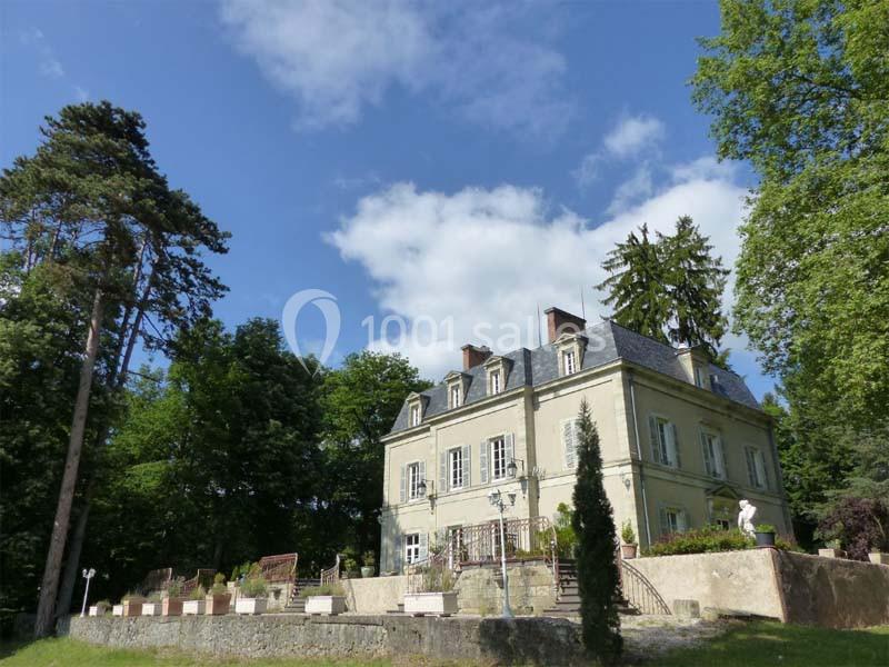 Manoir ancien entouré d'arbres, avec une terrasse en pierre et un ciel bleu dégagé en arrière-plan.