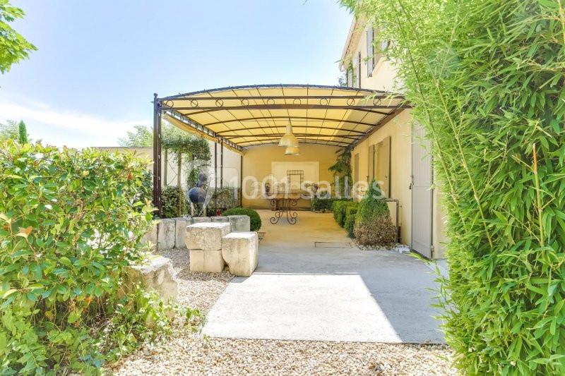 Terrasse couverte avec structure métallique, entourée de végétation, donnant sur une maison beige.