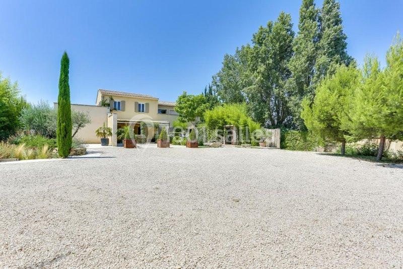 Maison avec façade beige entourée de végétation, vue depuis une grande cour en gravier sous un ciel bleu.