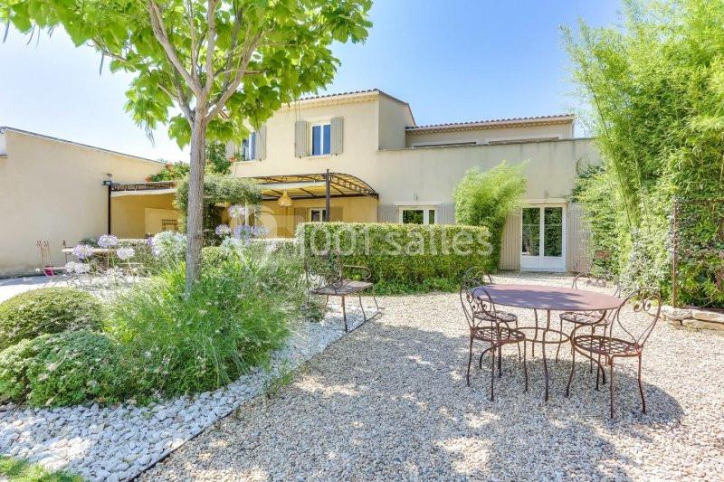 Jardin avec mobilier en fer forgé, gravier, végétation luxuriante et maison beige à deux étages en arrière-plan.