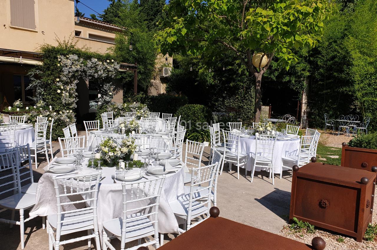Tables rondes dressées avec nappes blanches et chaises blanches dans un jardin, entourées de verdure et de fleurs.