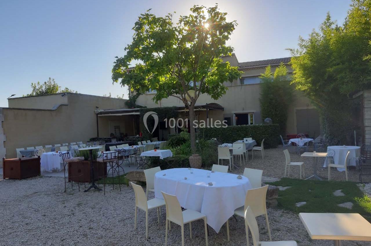 Terrasse extérieure avec tables et chaises blanches disposées sur du gravier, entourée de verdure et éclairée par le soleil.