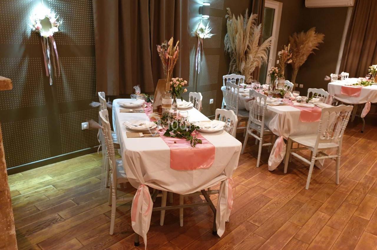 Salle décorée pour un événement avec tables dressées, nappes blanches, chemins de table roses et arrangements floraux.