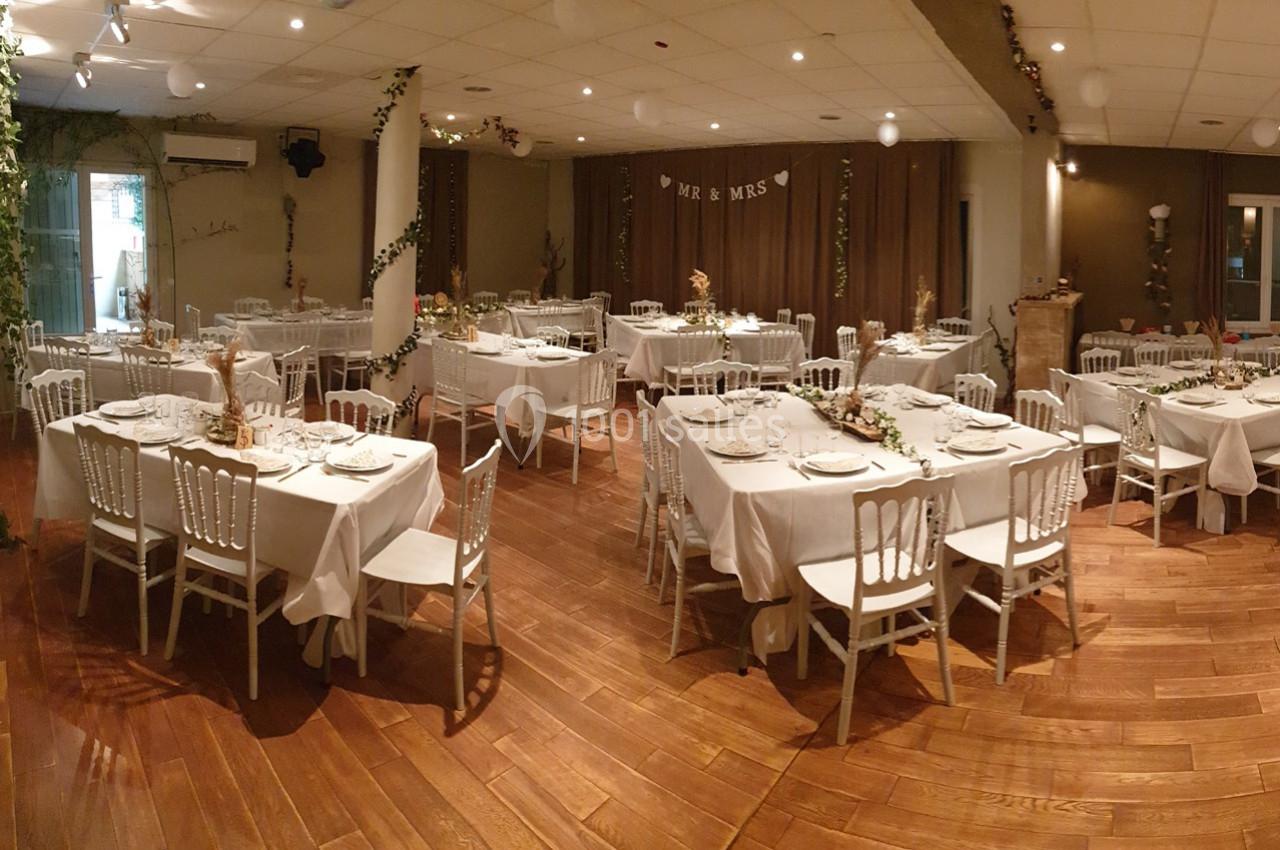 Salle de réception décorée pour un mariage, avec tables dressées, nappes blanches et chaises alignées sur un parquet en bois.