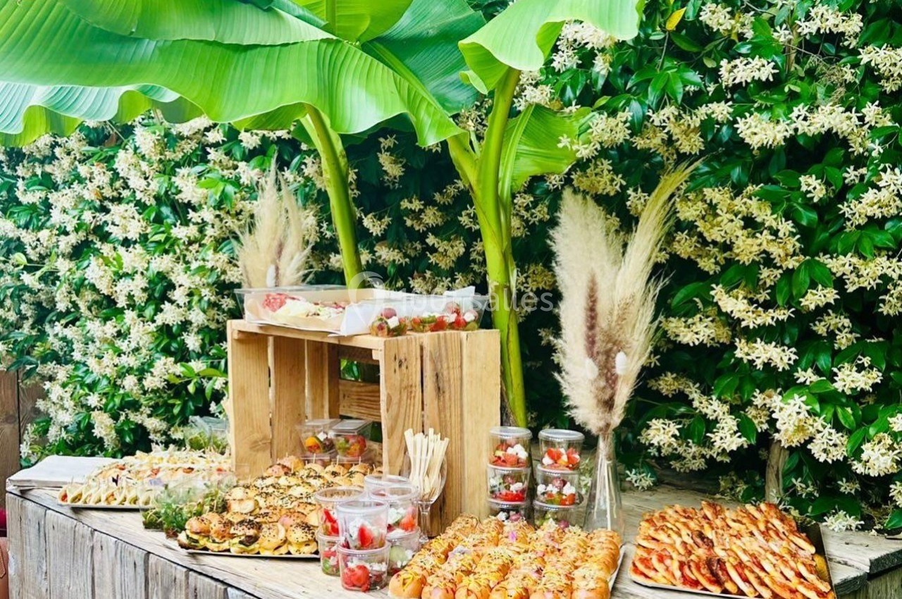 Buffet en plein air avec viennoiseries, brochettes, verrines et décorations végétales devant un mur fleuri.